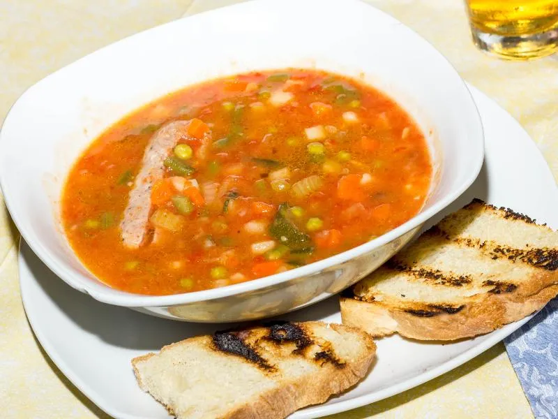 A bowl of red vegetable soup with celery, carrots, peas and pale beans, with two slices of grilled bread alongside