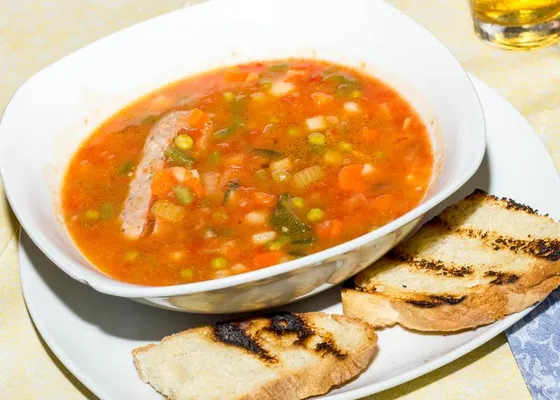 A bowl of red vegetable soup with celery, carrots, peas and pale beans, with two slices of grilled bread alongside