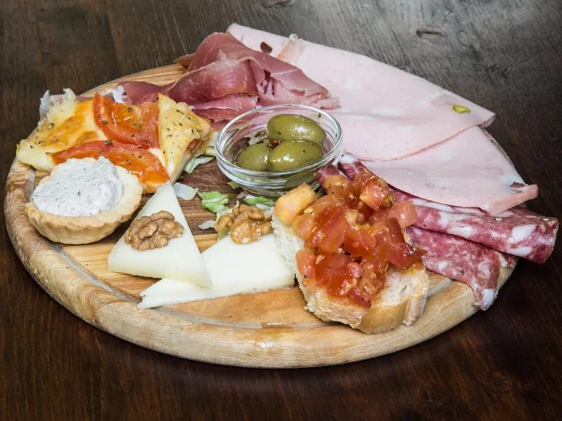 A wooden board of sliced cured meats, cheese, olives, tomato crostini and other small prepared bites