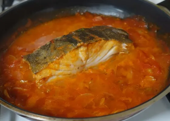 A thick skin-on cod portion resting in red tomato sauce with softened onion slices in a dark skillet