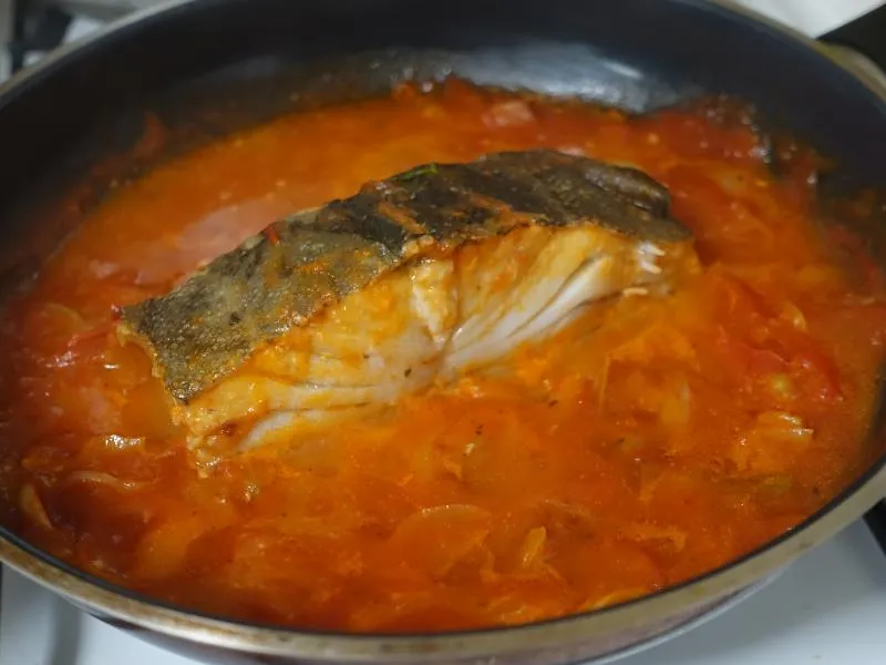 A thick skin-on cod portion resting in red tomato sauce with softened onion slices in a dark skillet