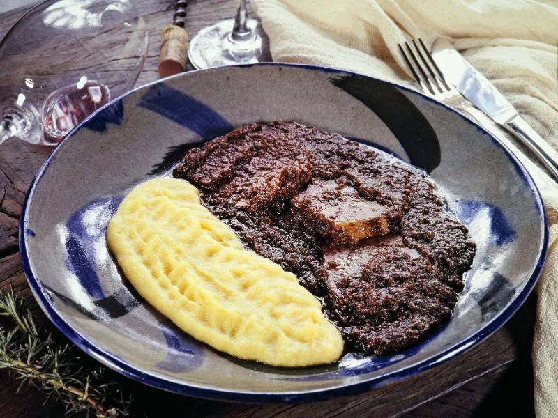 Slices of dark wine-braised beef covered with a thick sauce beside yellow polenta on a blue and white plate