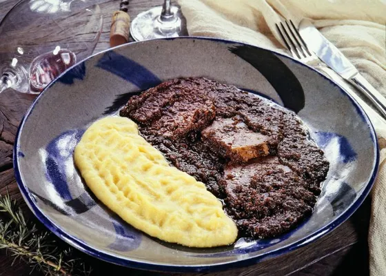 Slices of dark wine-braised beef covered with a thick sauce beside yellow polenta on a blue and white plate