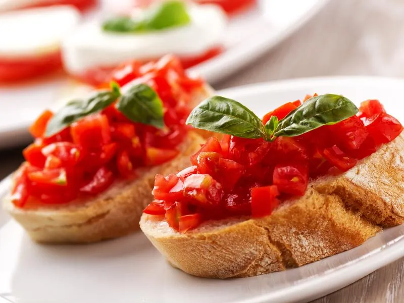 Thick slices of toasted bread topped with chopped red tomatoes and fresh basil leaves on a white plate