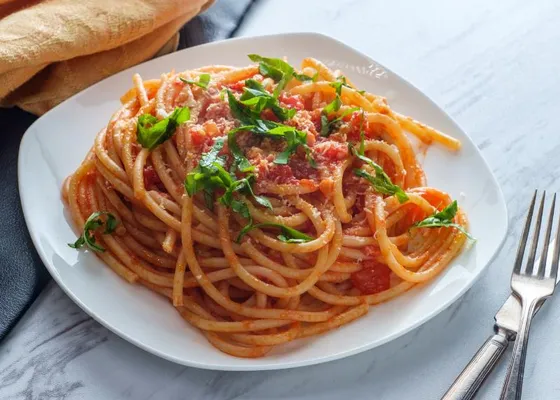 Long bucatini in red tomato sauce with pieces of cured pork, grated cheese and green herb leaves on a white plate