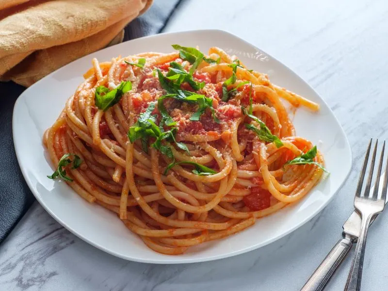 Long bucatini in red tomato sauce with pieces of cured pork, grated cheese and green herb leaves on a white plate