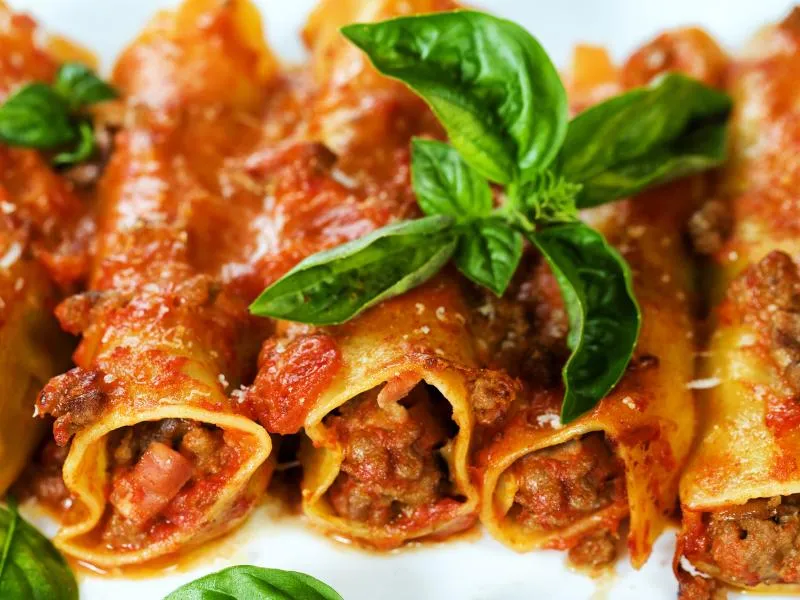 Meat-filled pasta rolls with red tomato sauce, grated cheese and broad basil leaves on a white plate