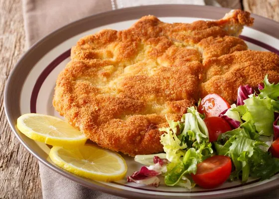 A broad golden breaded veal cutlet with a visible rib bone, lemon slices and mixed salad on a white plate