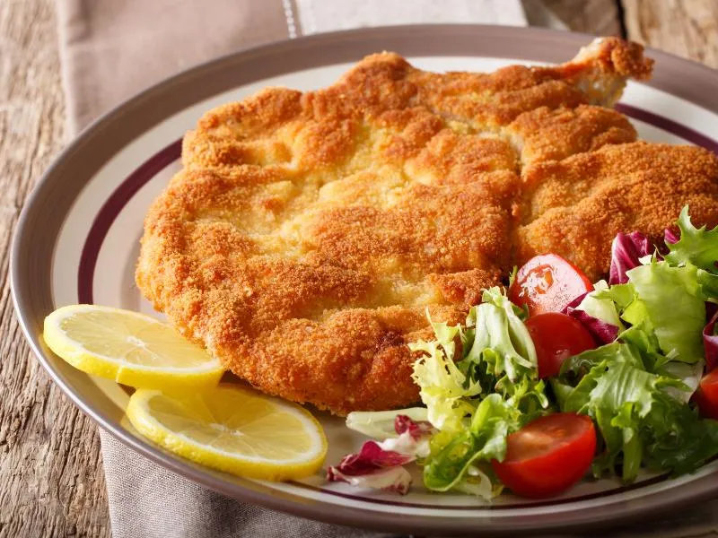 A broad golden breaded veal cutlet with a visible rib bone, lemon slices and mixed salad on a white plate