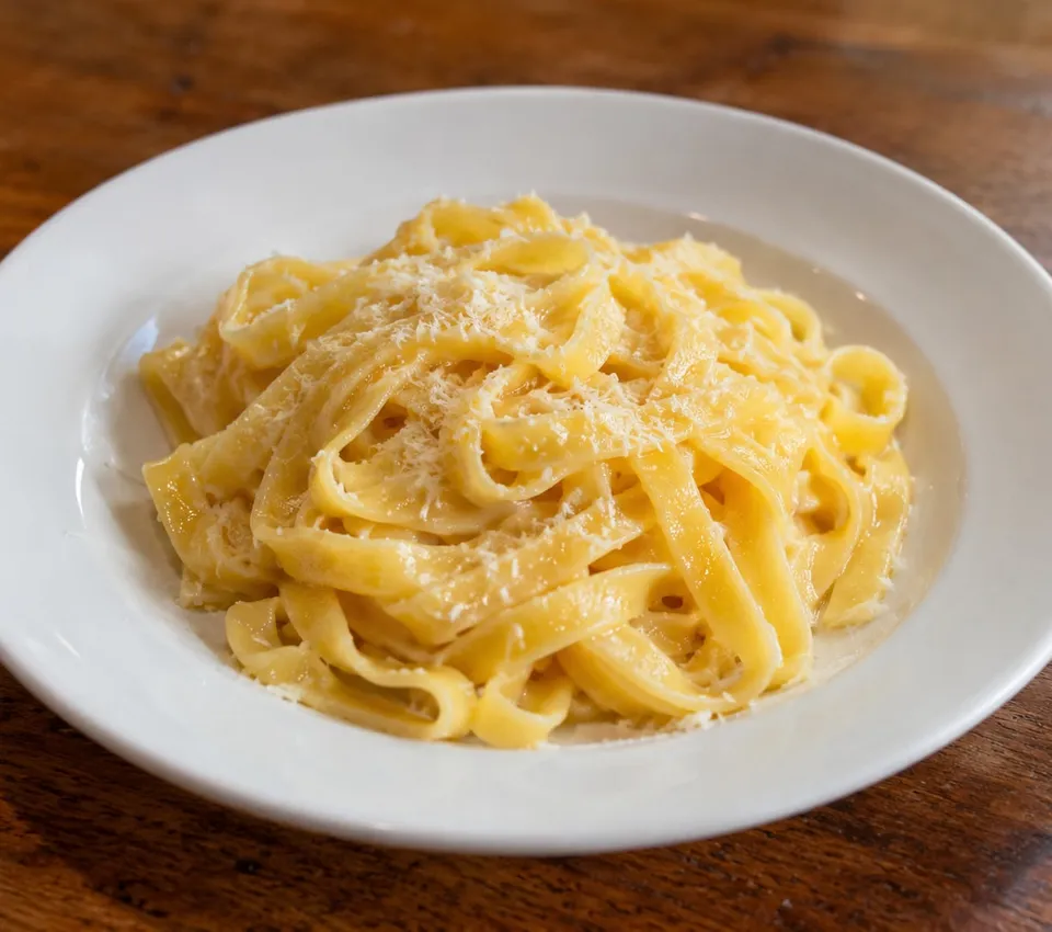 Golden fettuccine ribbons with a glossy butter-and-cheese coating and grated Parmigiano on a white plate