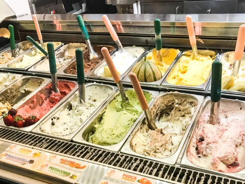 A gelateria display of metal tubs containing several colors and flavors of gelato, with serving paddles