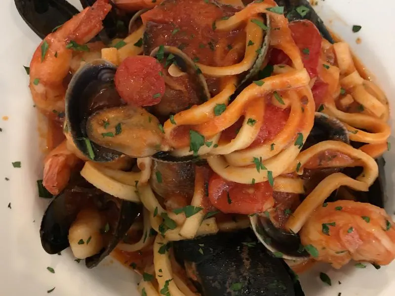 Long flat pasta with mussels, shrimp, tomato pieces and chopped parsley on a white plate