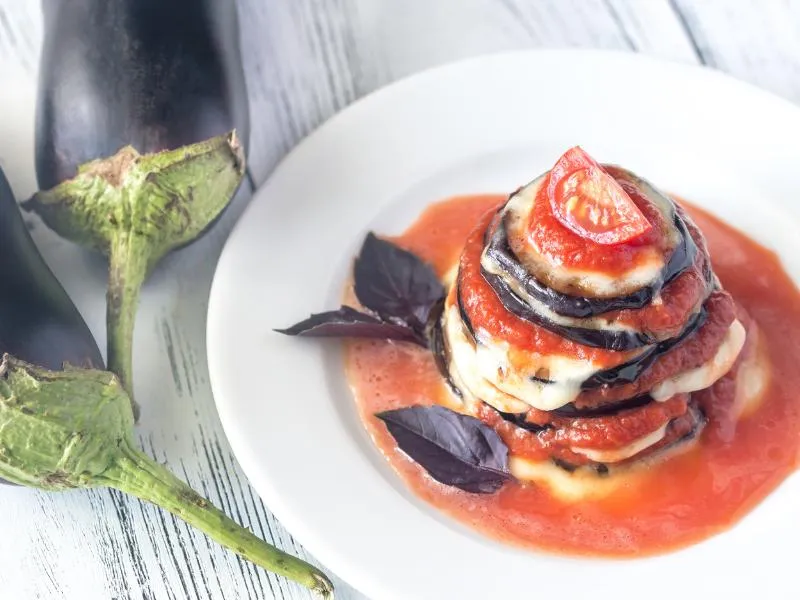 An individual stack of eggplant rounds, melted cheese and tomato sauce, topped with a tomato slice and purple basil leaves