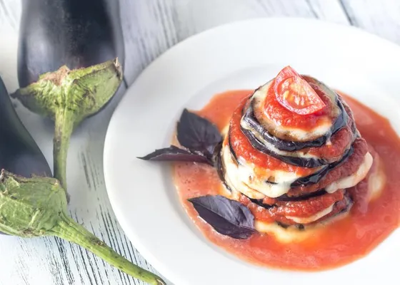 An individual stack of eggplant rounds, melted cheese and tomato sauce, topped with a tomato slice and purple basil leaves