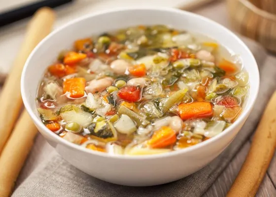 A bowl of vegetable soup with white beans, carrots, potatoes and green vegetables in a light tomato broth