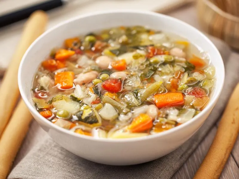 A bowl of vegetable soup with white beans, carrots, potatoes and green vegetables in a light tomato broth
