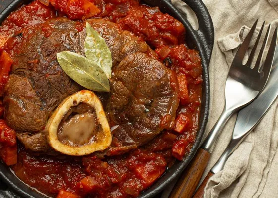 A braised shank with a central marrow bone in red tomato sauce, topped with bay leaves in a dark pan