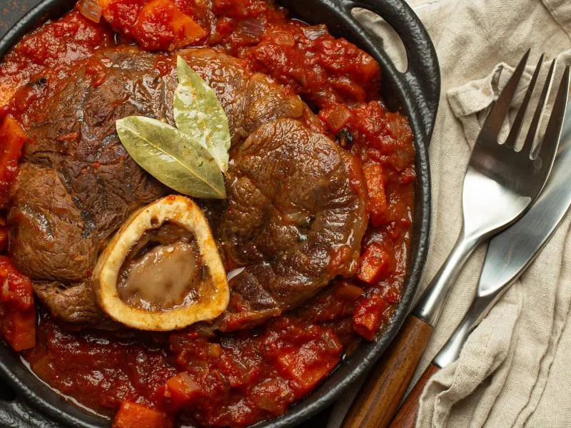 A braised shank with a central marrow bone in red tomato sauce, topped with bay leaves in a dark pan