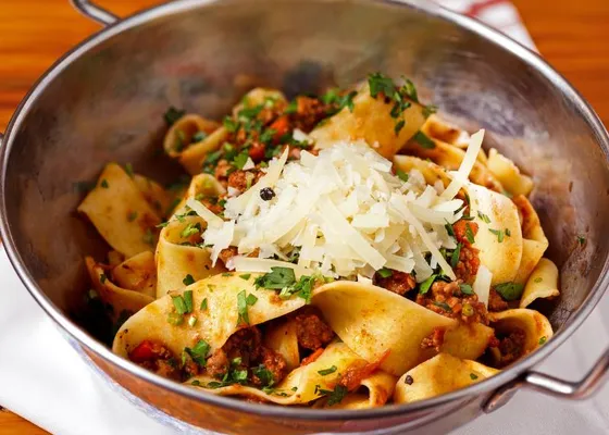 Wide pasta ribbons with red-brown meat sauce, chopped herbs and shaved cheese in a small metal pan