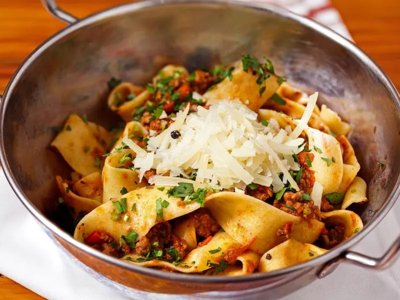 Wide pasta ribbons with red-brown meat sauce, chopped herbs and shaved cheese in a small metal pan