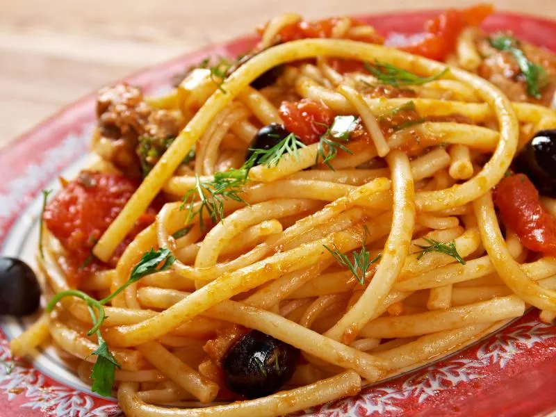 Long pasta in a chunky tomato sauce with dark olives and a mixed fresh-herb garnish on a red patterned plate