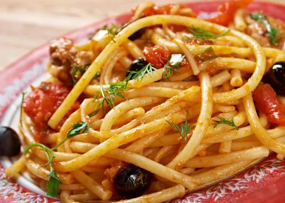 Long pasta in a chunky tomato sauce with dark olives and a mixed fresh-herb garnish on a red patterned plate