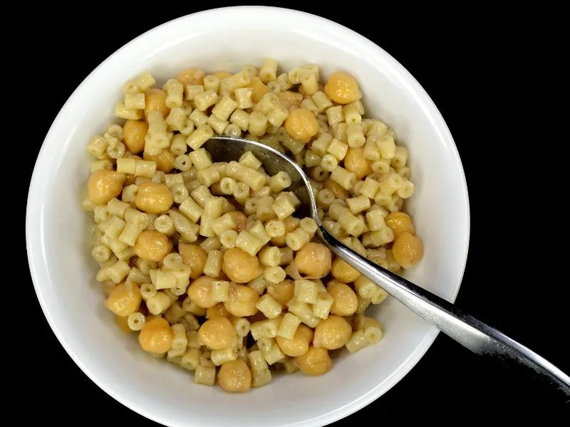A thick serving of small tubular pasta and chickpeas in a white bowl with a spoon, against a dark background