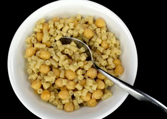 A thick serving of small tubular pasta and chickpeas in a white bowl with a spoon, against a dark background