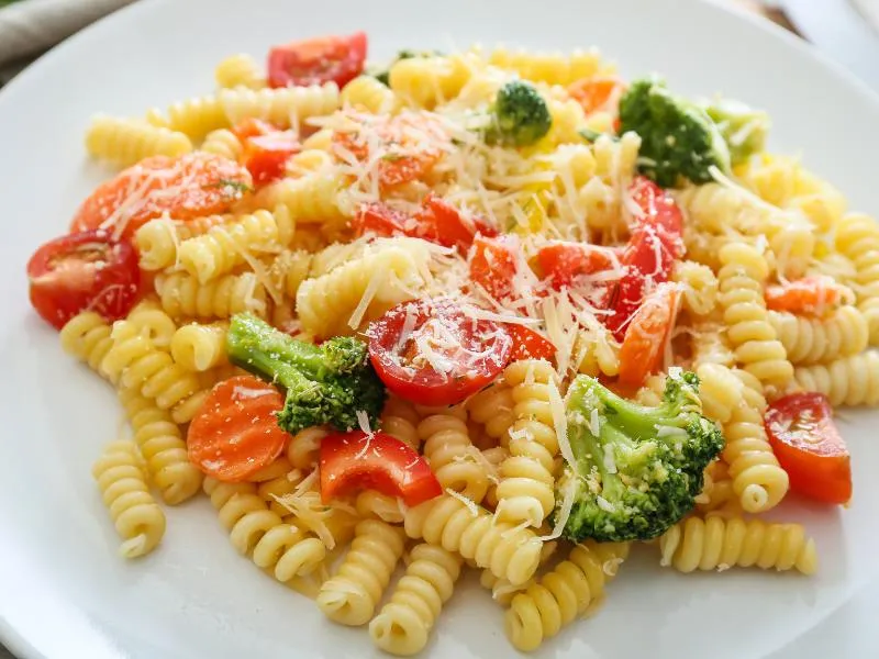 Spiral pasta with broccoli, carrot slices, red pepper, tomato halves and grated cheese on a white plate