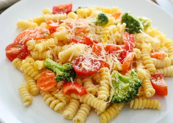 Spiral pasta with broccoli, carrot slices, red pepper, tomato halves and grated cheese on a white plate