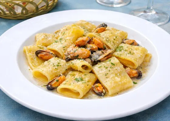 Large paccheri pasta tubes with shelled orange mussels, grated cheese and chopped green herbs on a white plate