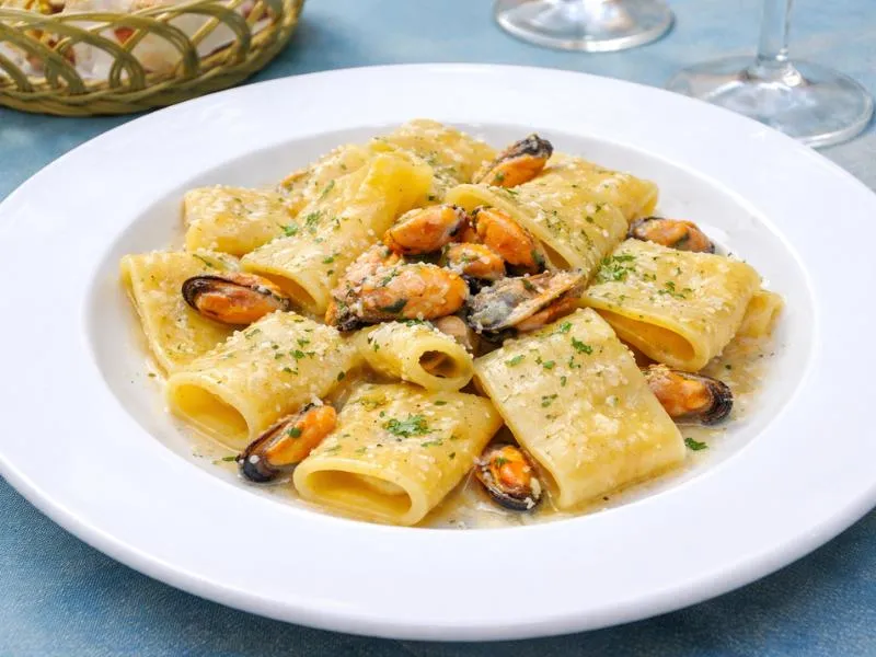 Large paccheri pasta tubes with shelled orange mussels, grated cheese and chopped green herbs on a white plate