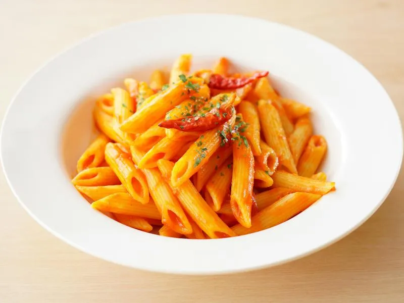 Ridged penne coated in red tomato sauce, topped with whole red chilies and chopped green parsley on a white plate