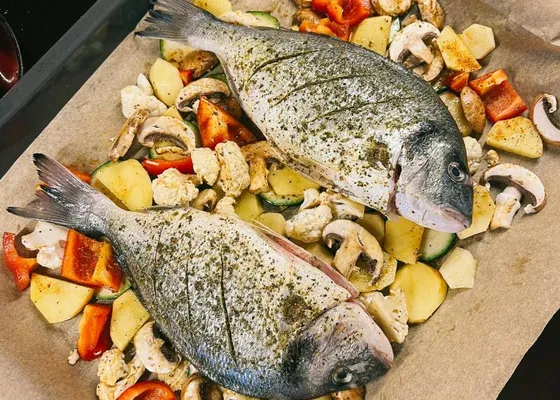 Two whole sea bream on a lined baking tray among potatoes, carrots, zucchini, mushrooms and cauliflower