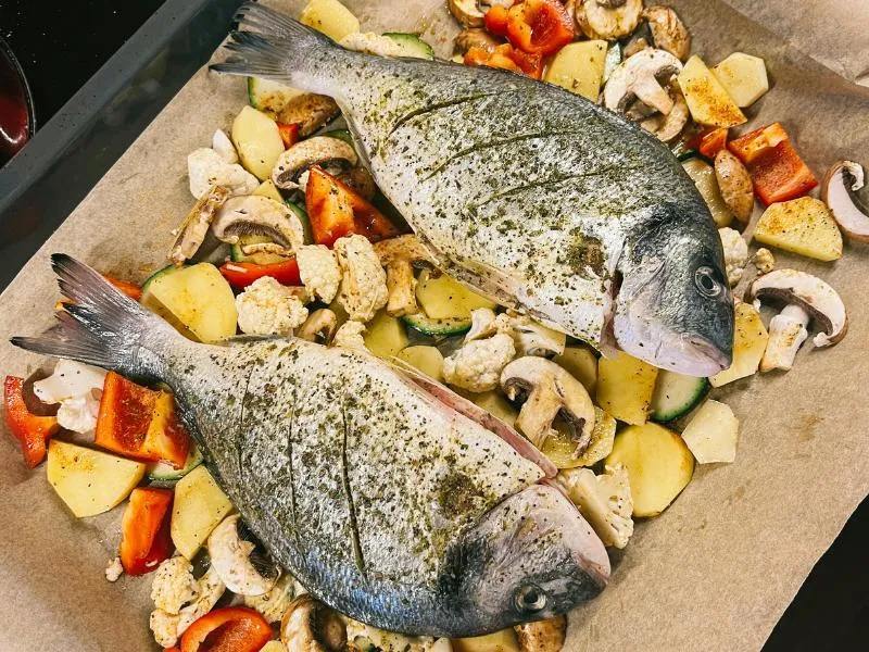 Two whole sea bream on a lined baking tray among potatoes, carrots, zucchini, mushrooms and cauliflower