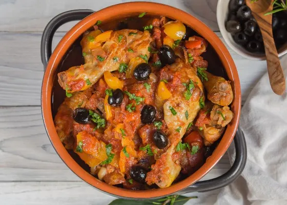 Chicken pieces in red tomato sauce with black olives, yellow pepper pieces and chopped herbs in a terracotta casserole