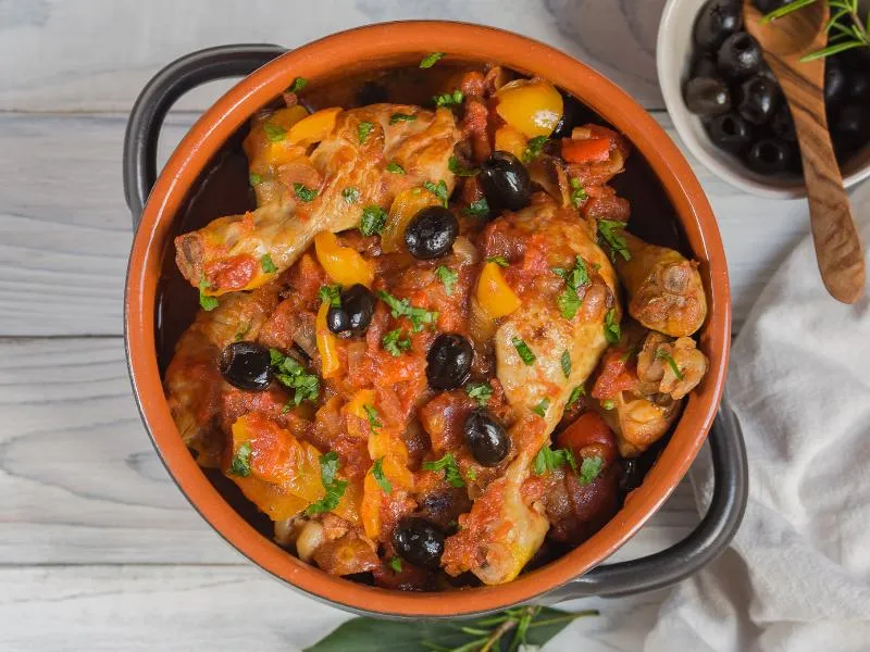 Chicken pieces in red tomato sauce with black olives, yellow pepper pieces and chopped herbs in a terracotta casserole