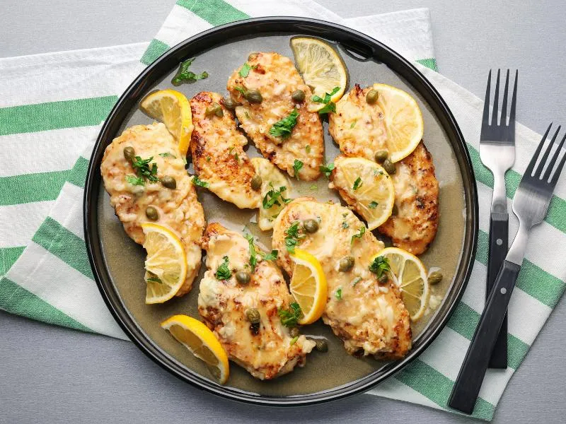Golden chicken cutlets topped with capers, lemon slices and chopped parsley on a dark plate