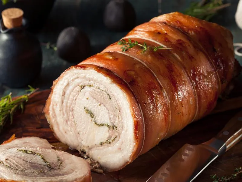 Sliced rolled pork roast with golden brown skin, herb-speckled spirals and a lean center on a wooden board