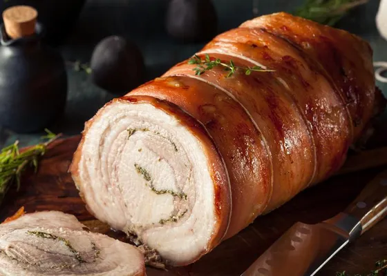 Sliced rolled pork roast with golden brown skin, herb-speckled spirals and a lean center on a wooden board