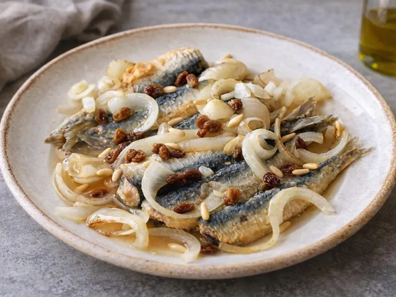 Opened sardine fillets layered with pale onion rings, raisins and pine nuts on a ceramic plate