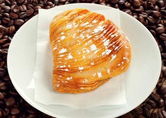 A golden shell-shaped pastry with many crisp ridges and a dusting of powdered sugar on a white plate