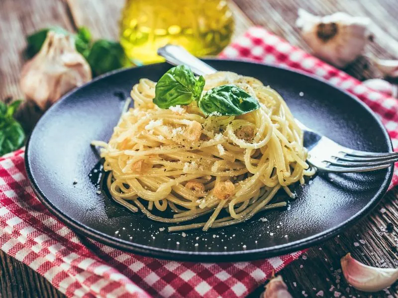 Spaghetti with sliced golden garlic and a dusting of grated cheese, topped with basil leaves on a dark plate