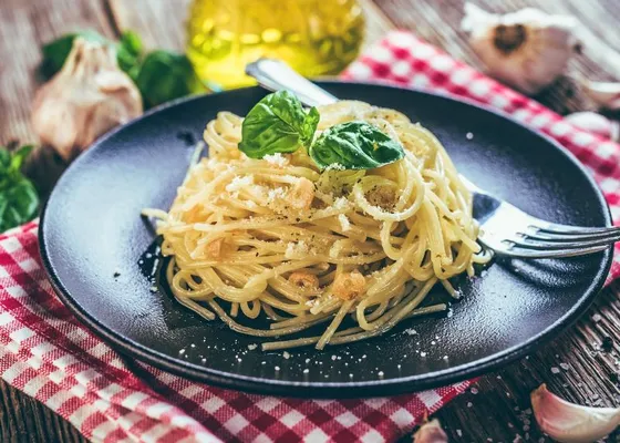 Spaghetti with sliced golden garlic and a dusting of grated cheese, topped with basil leaves on a dark plate