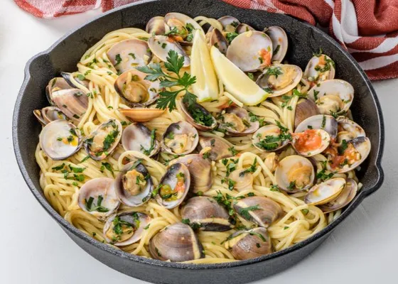 Spaghetti with opened clams, parsley, a few red tomato pieces and lemon wedges in a dark skillet