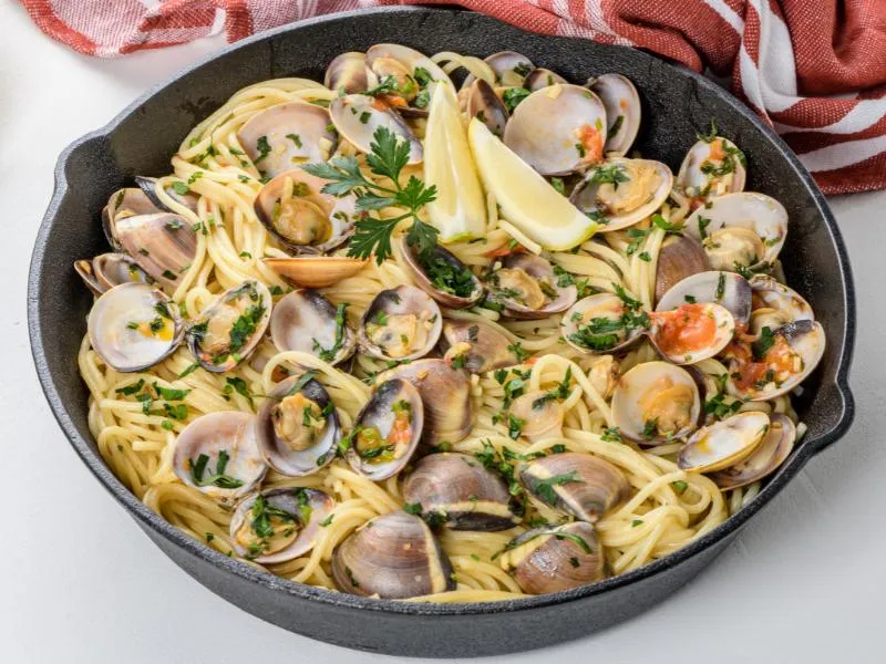 Spaghetti with opened clams, parsley, a few red tomato pieces and lemon wedges in a dark skillet