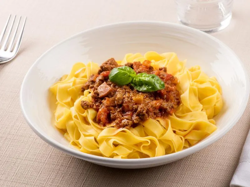 Wide golden egg-pasta ribbons topped with brown-red meat sauce and a basil sprig on a white plate
