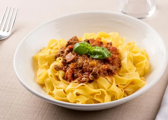 Wide golden egg-pasta ribbons topped with brown-red meat sauce and a basil sprig on a white plate