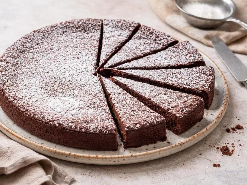 A round dark chocolate cake cut into wedges and dusted with powdered sugar on a ceramic plate