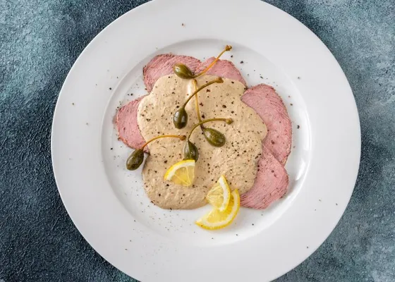 Thin pale-pink veal slices under a creamy tuna sauce with caper berries, lemon wedges and black pepper on a white plate